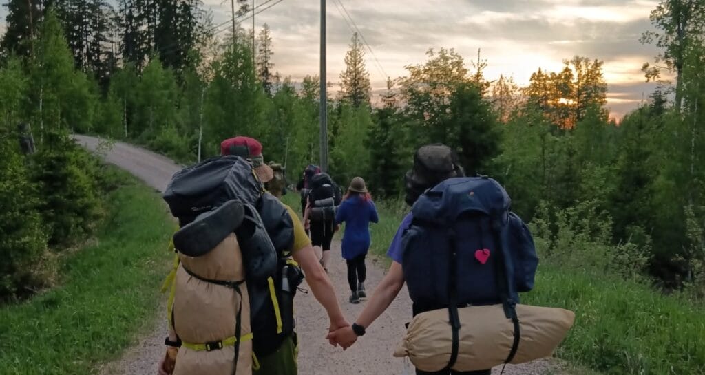 Vaeltajia rinkat selässä polulla, kuvattuna takaapäin kulkemassa eteenpäin. Kaksi henkilöä pitävät toisiaan kädestä kiinni kävellessä.